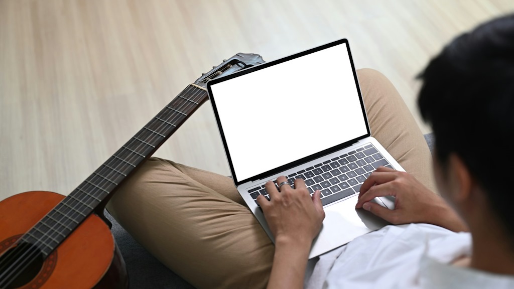 Un homme tape sur son ordinateur avec une guitare sur le genou gauche.