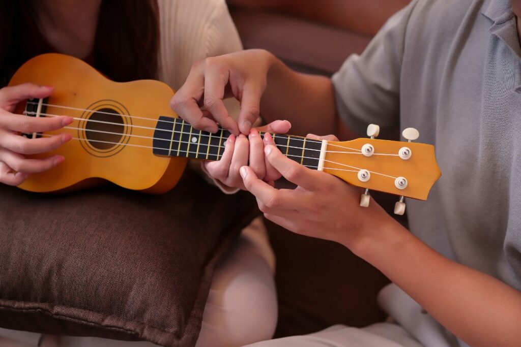 Deux mains guidant le placement des doigts sur un ukulélé soprano lors d'un cours.