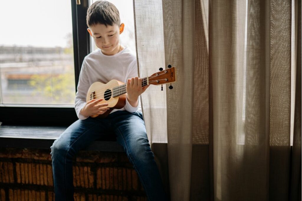 Jeune garçon souriant en jouant du ukulélé près d'une fenêtre.