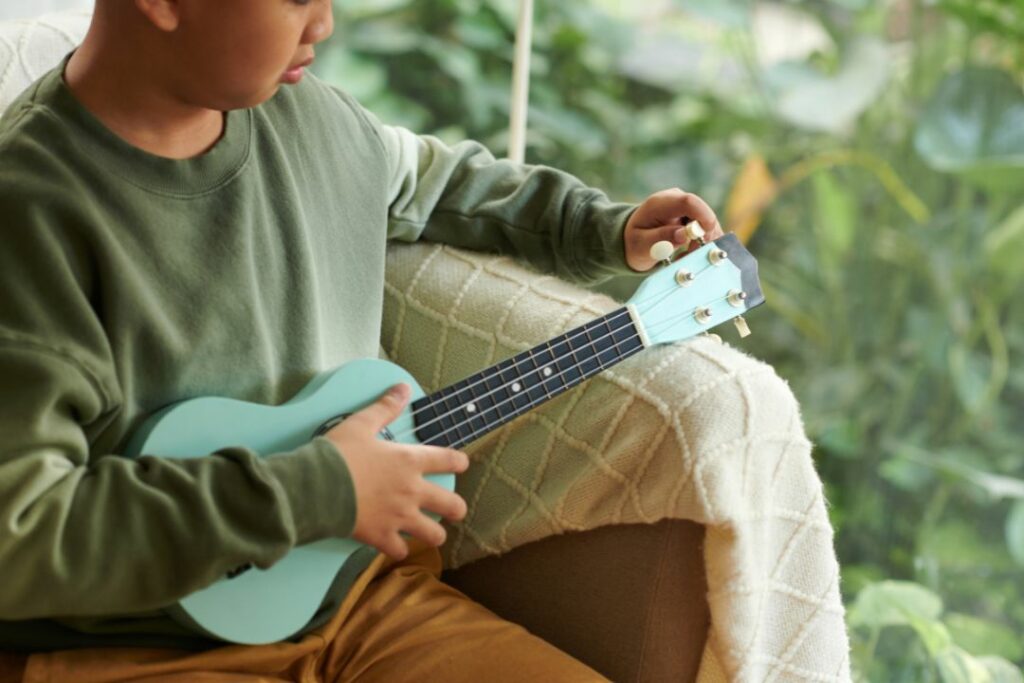 Jeune garçon ajustant les mécaniques d'un ukulélé bleu ciel sur un canapé.