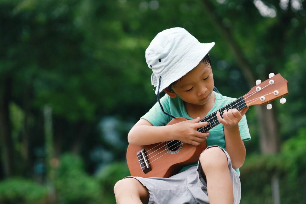 Jeune enfant avec un chapeau apprenant à jouer du ukulélé soprano dans un parc.
