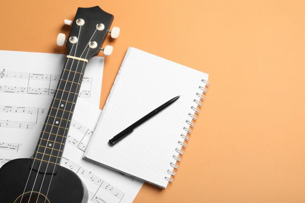 Ukulélé noir posé sur une partition à côté d'un cahier et d'un stylo.